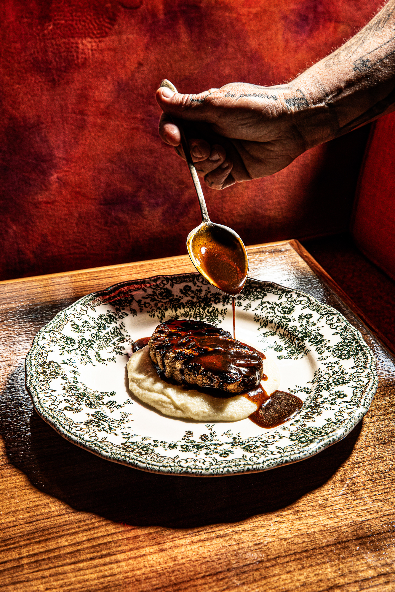 Hand spooning sauce over steak on a toile plate in a red booth