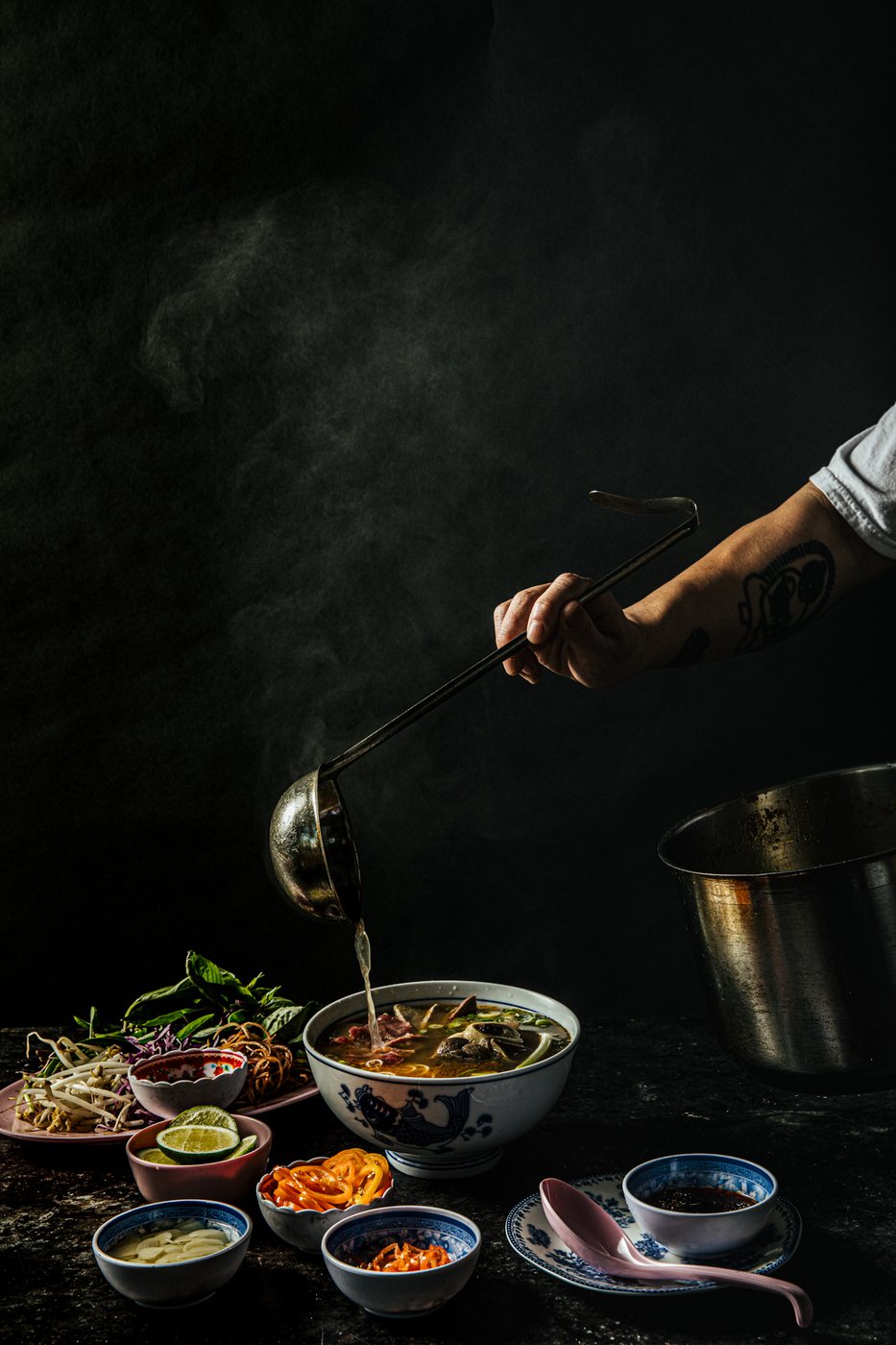 Steam rising from a bowl of pho as broth is ladled in, dark ground